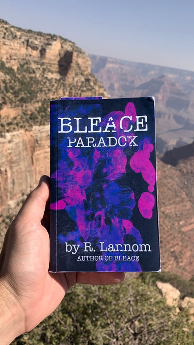 Photograph of a Bleace Paradox soft cover novel that has splattered pink and purple paint on it with white text that says ‘Bleace Paradox by R. Lannom Author of Bleace’ on the front cover that is in the foreground with the Grand Canyon, Arizona in the background.