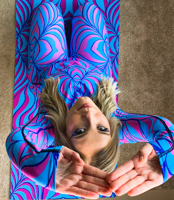 Photograph of a model wearing a Bleace blue and pink yoga leggings and rash guard in the foreground with a matching yoga mat in the background.