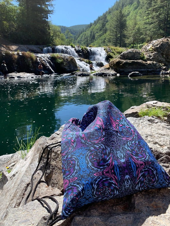 Photograph of a Bleace psychedelic print drawstring bag in the foreground that has pink, turquoise and purple swirls on it with a waterfall in Washington state in the background.