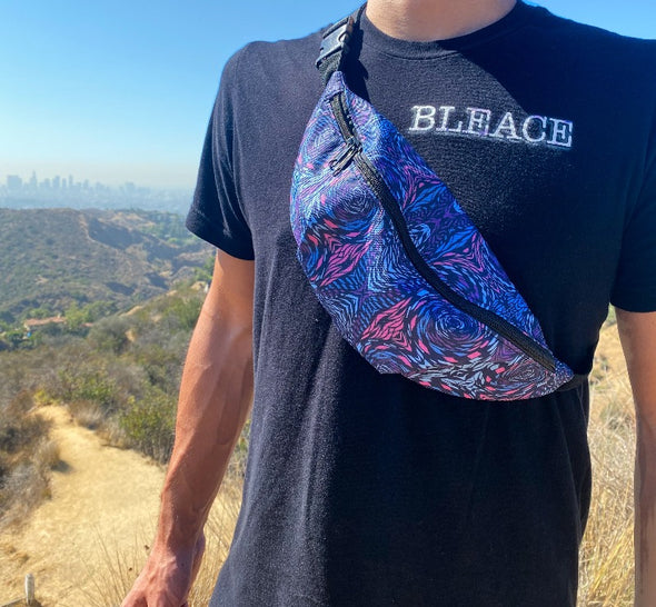 Photograph of a Bleace psychedelic print fanny pack being worn over the shoulder of a male model who is wearing a black Bleace shirt standing in the foreground, with the Los Angeles cityscape in the background. 