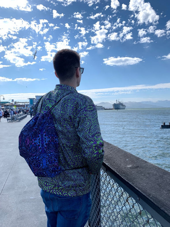 Photograph of a Bleace psychedelic print drawstring bag in the foreground being word by a male model staring off towards the Pacific ocean wearing a green Bleace bomber jacket in San Francisco, California.