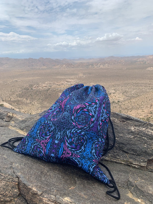 Photograph of a Bleace psychedelic print drawstring bag in the foreground that has pink, turquoise and purple swirls on it with mountains at the Joshua Tree National Park in California in the background.